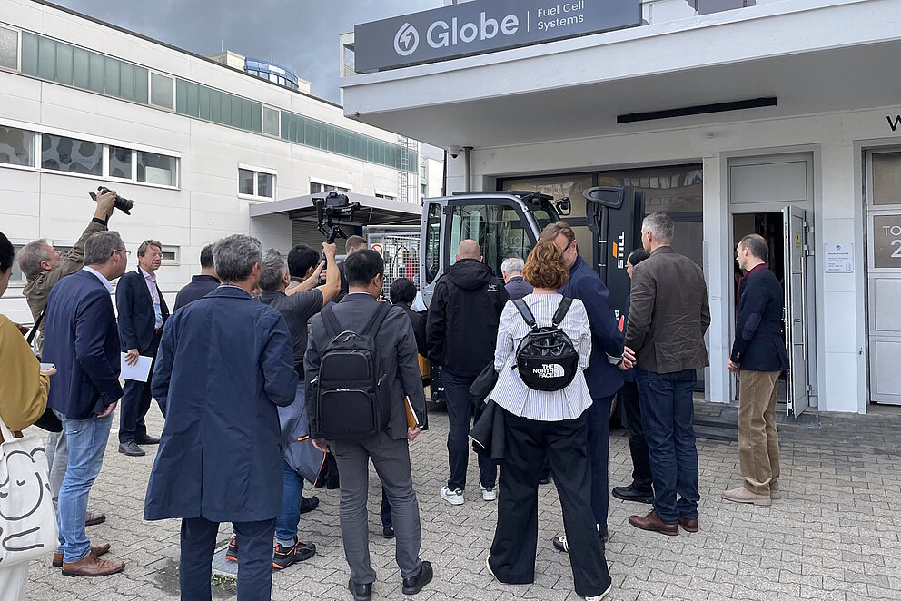 Eine Gruppe Menschen steht vor dem Eingang eines Unternehmens und betrachtet ein dort geparktes Nutzfahrzeug.