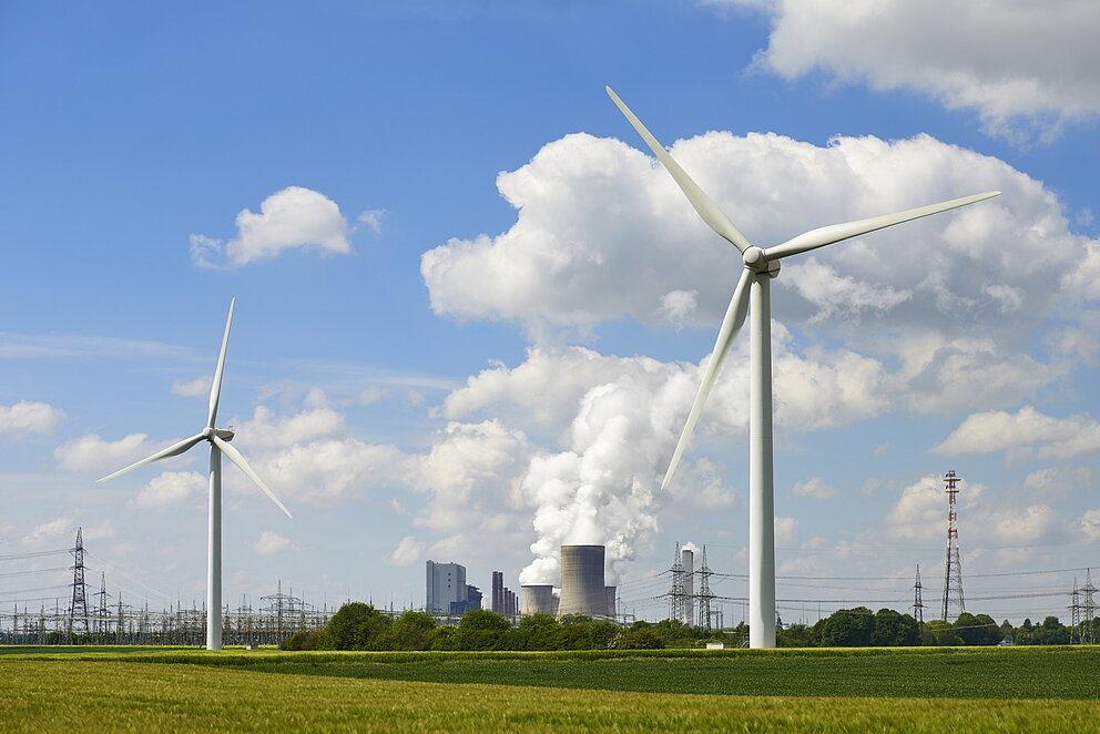 Sommerliche Landschaftsaufnahme mit Windrädern im Vordergrund und einem Kraftwerk mit rauchenden Türmen im Hintergrund.