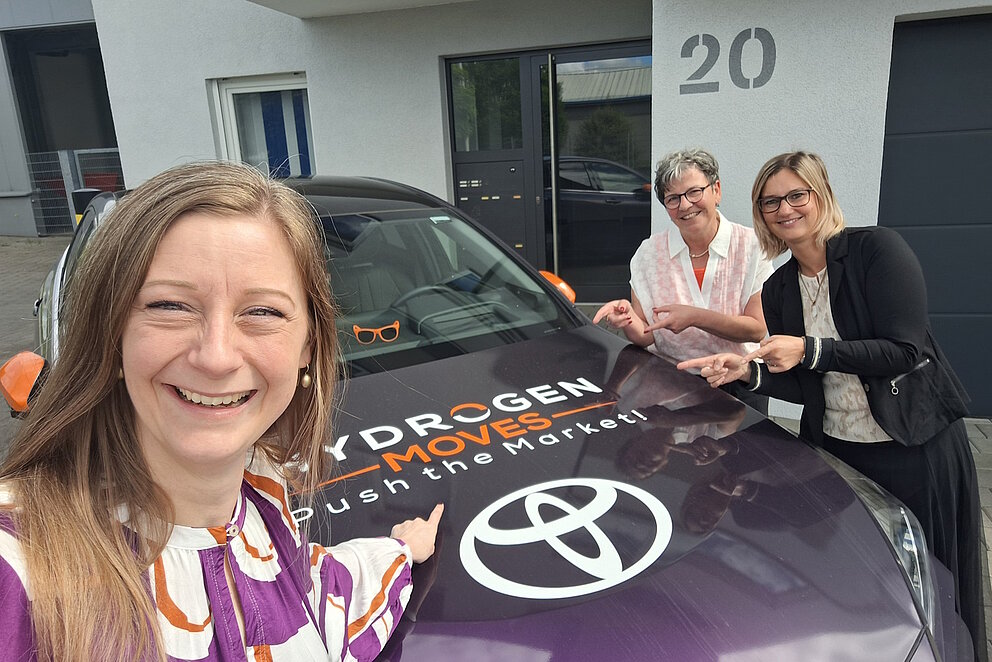 Die drei Mitarbeiterinnen bei Hydrogen Moves posieren vor einem Wasserstoff-Auto mit aufgedrucktem Logo von Hydrogen Moves.