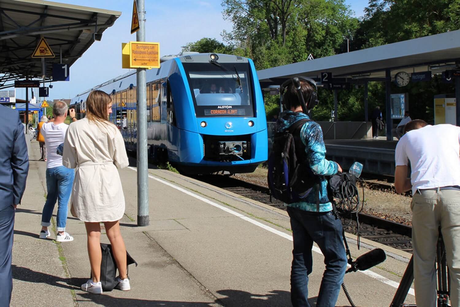 An einem Bahngleis auf dem wartende Menschen stehen fährt ein blauer Zug ein.