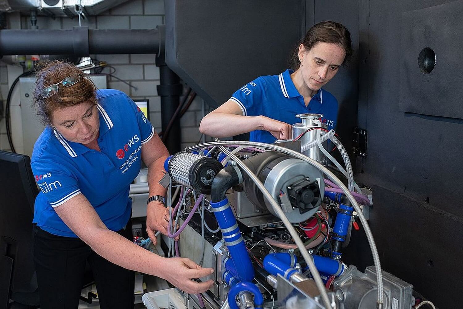 Zwei Frauen mit blauen T-Shirts beugen sich über ein Modell einer Brennstoffzelle mit vielen Schläuchen.