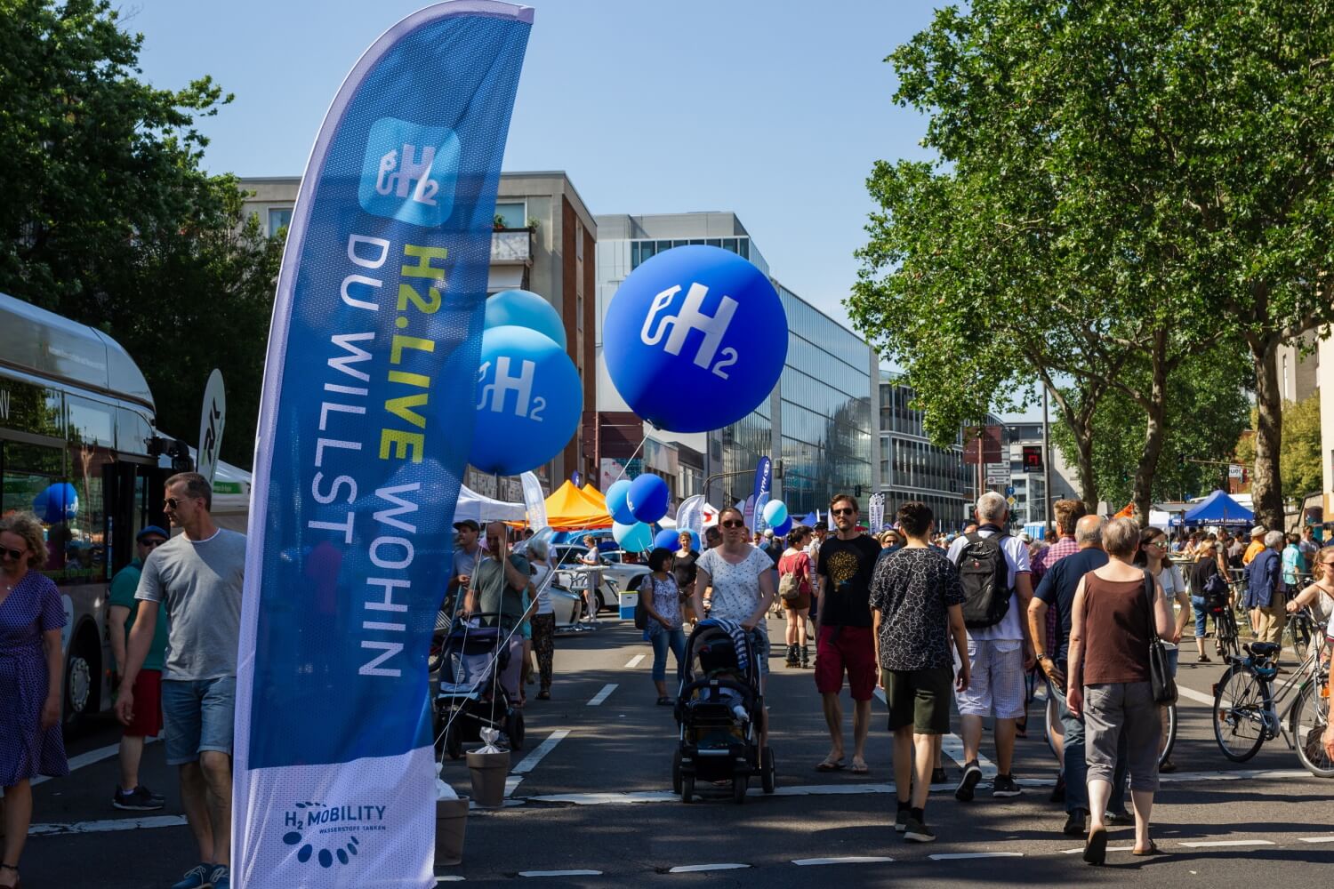 Auf einer von Bäumen und Häusern gesäumten Straße gehen viele Menschen an in Blau geschmückten Ständen und Ballons vorbeit; dabei sind die Schriftzüge H2 und Wasserstoff zu sehen.