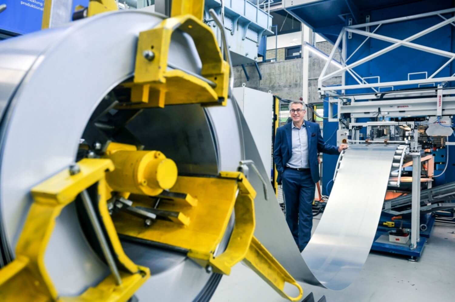 In einer hellen Fertigungshalle läuft ein Mann im Hintergrund im Anzug auf eine große Rolle von silbernem Metallstreifen im Vordergrund zu.