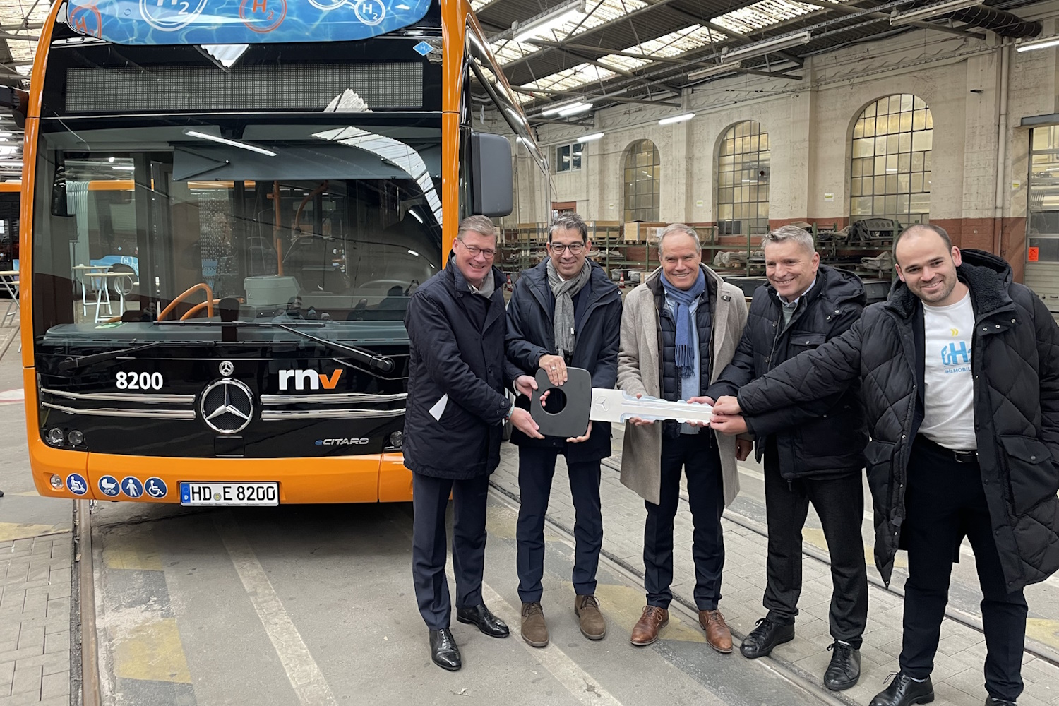 Eine gruppe Männder steht vor Wasserstoffbus in einer Halle und halten einen überdimensionierten Schlüssel in den Händen.